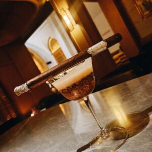 Premium cigar resting on cocktail glass with amber liquid in elegant lounge setting with warm lighting